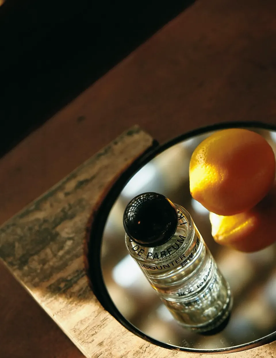 Perfume bottle and yellow fruit on a reflective tray in natural light.